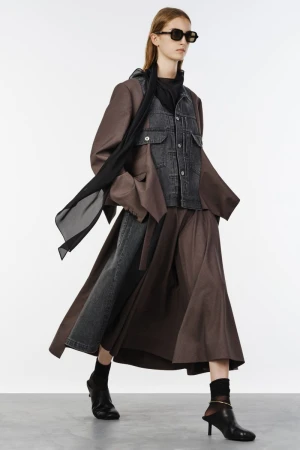 Fashion model walking in layered brown outerwear, grey denim jacket, dark sunglasses, and black heeled mules on white studio seamless.
