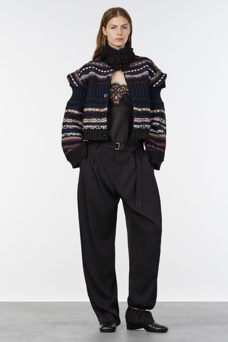 Model wearing a patterned cropped knit jacket over a lace-trim top and pleated wide-leg black trousers against a white studio backdrop.