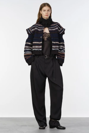 Model wearing a patterned cropped knit jacket over a lace-trim top and pleated wide-leg black trousers against a white studio backdrop.