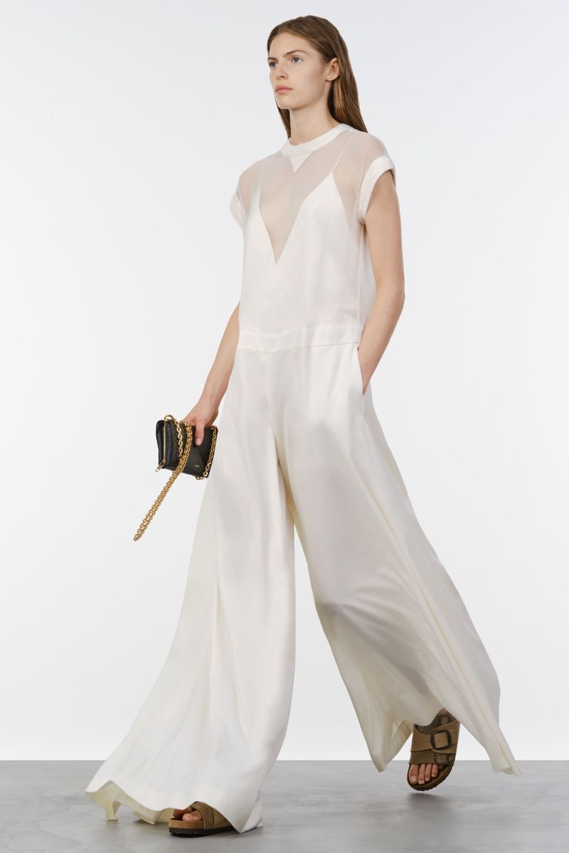 Model walking in a flowing white wide-leg jumpsuit holding a black chain clutch against a bright white studio backdrop.