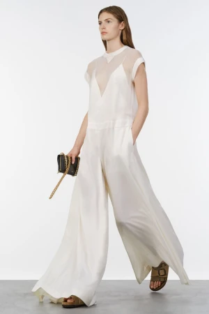 Model walking in a flowing white wide-leg jumpsuit holding a black chain clutch against a bright white studio backdrop.