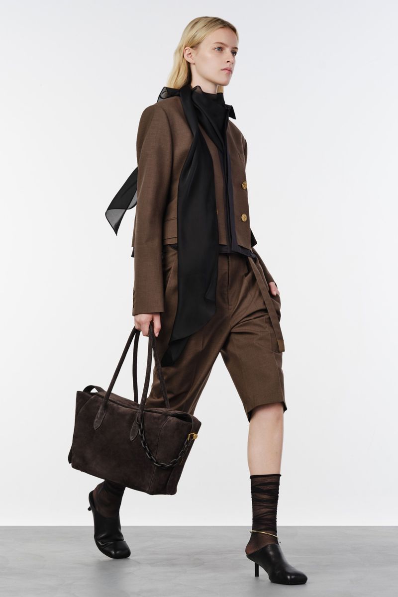 Model walking in a brown tailored jacket, matching shorts, black scarf, and kitten heels holding a large brown tote bag against a white background.