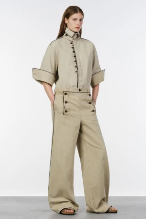 Model wearing a beige high-collar buttoned shirt and matching wide-leg trousers with dark piping on a white studio background.