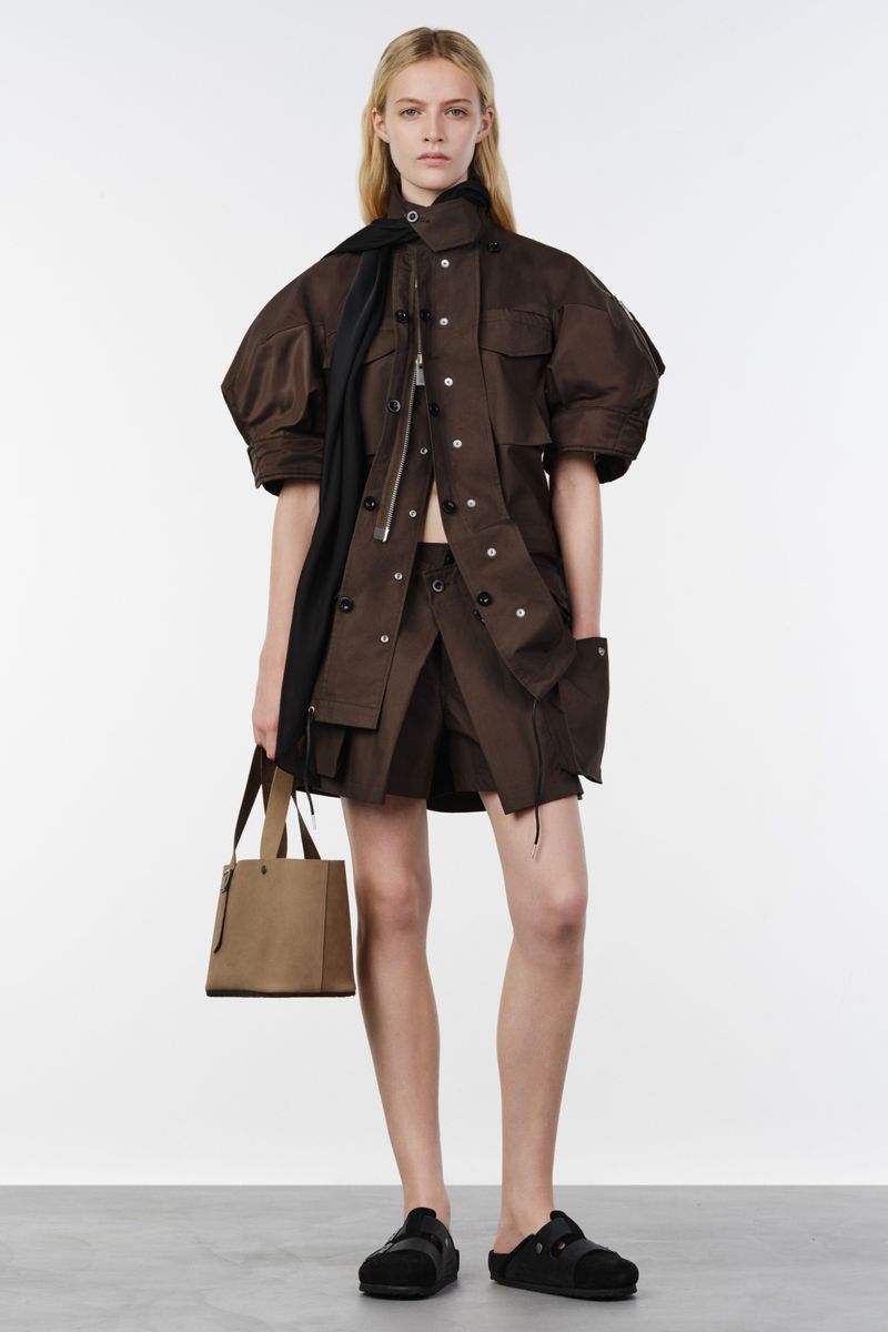 Model posing in a brown short-sleeve utility jacket, tailored shorts, and black slide sandals against a white backdrop.