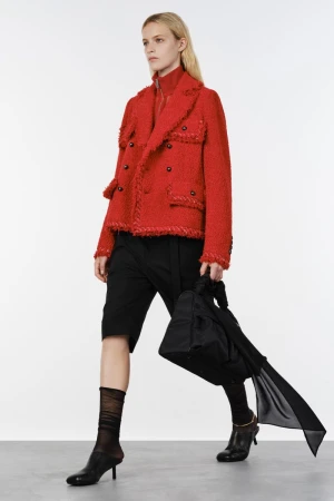 Model walking in a vibrant red textured jacket, black knee-length shorts, and black kitten heels against a white studio background.
