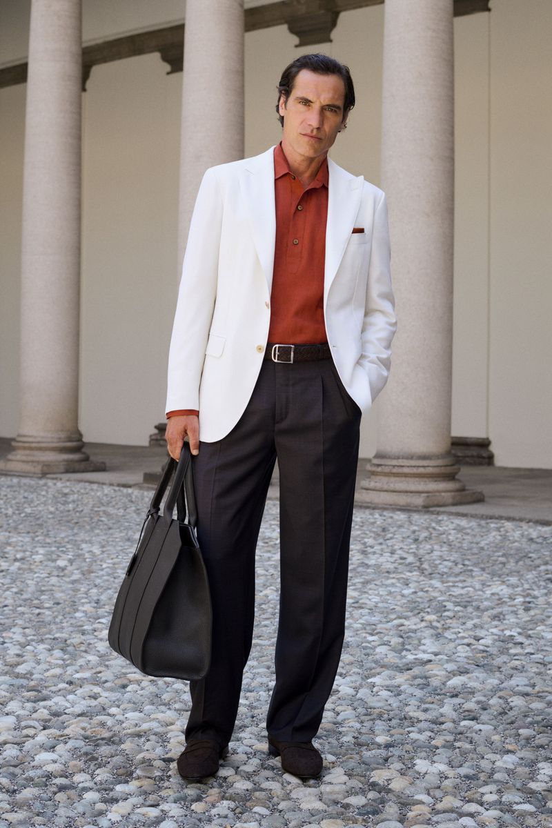 A man in a white blazer, terracotta polo shirt, and dark pleated trousers holding a large leather tote bag by stone columns.