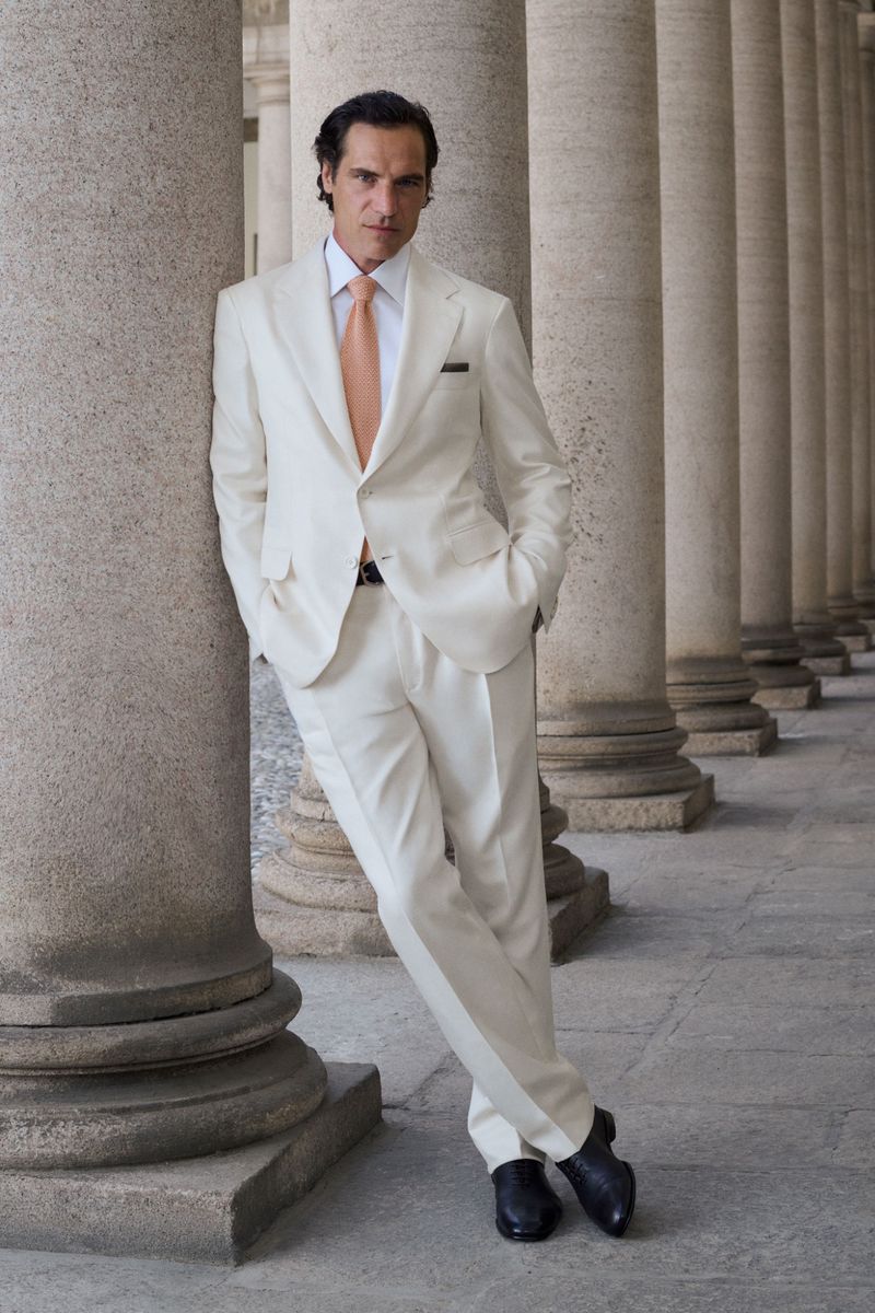 A man wearing an off-white tailored suit and peach tie leans against a classical stone column.