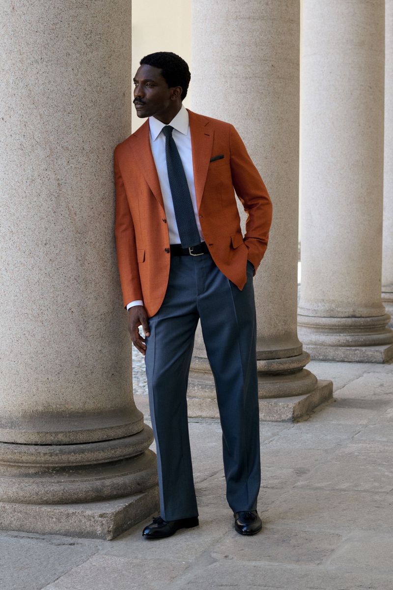Model Robbie Jr wearing a tailored burnt orange blazer and slate blue trousers leaning against a stone column for Brioni.