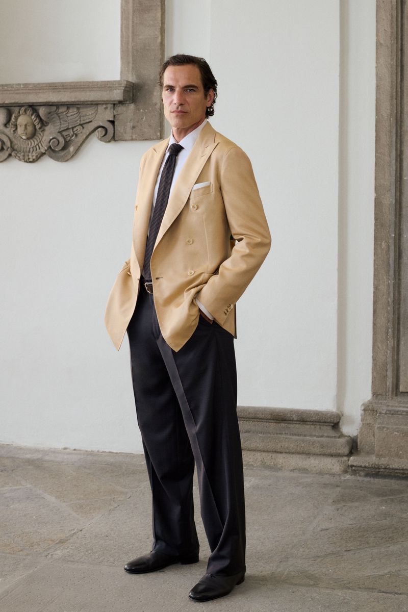 Man in a tan double-breasted blazer and dark tailored trousers standing in an architectural stone hallway.