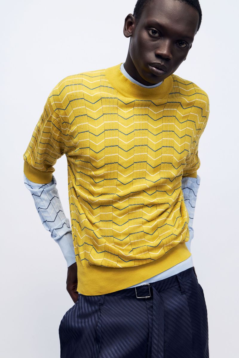 Model Bukwop Kir wearing a yellow chevron knit shirt over light blue sleeves and navy trousers against a white studio backdrop.