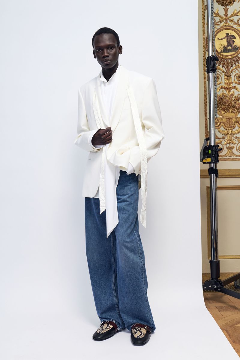 Model wearing a white blazer over blue jeans standing against a white backdrop with a lighting stand visible on the right.