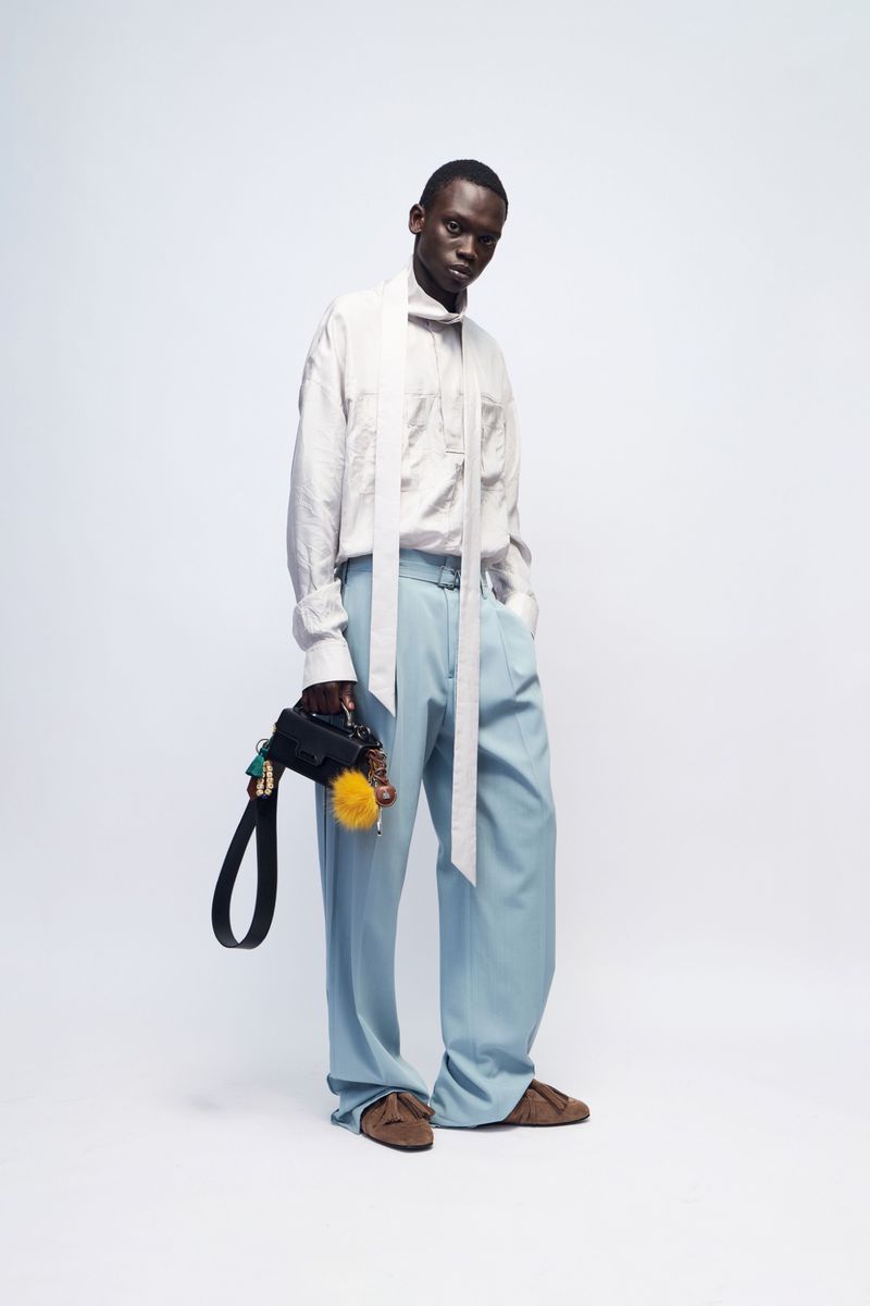 Model Bukwop Kir wearing a white scarf-collar shirt and wide-leg light blue trousers while holding a black mini bag on a white backdrop.