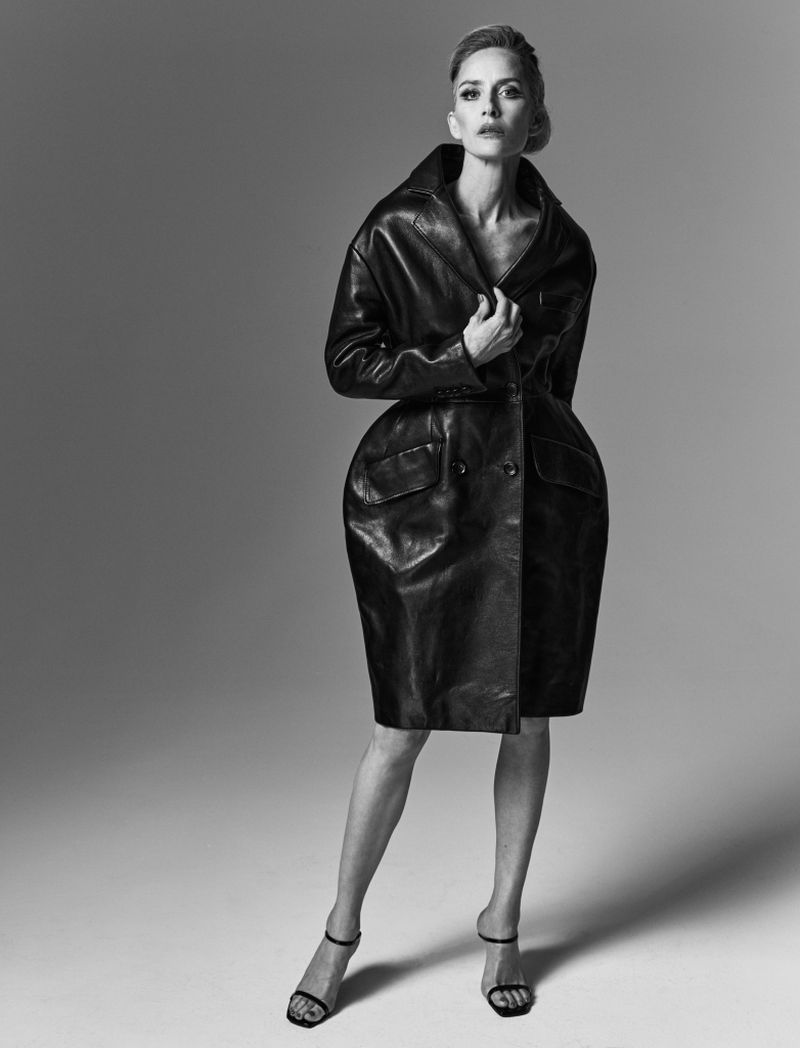 Black and white full shot of a blond woman in a black leather trench coat and open-toe heeled sandals in a grey studio.