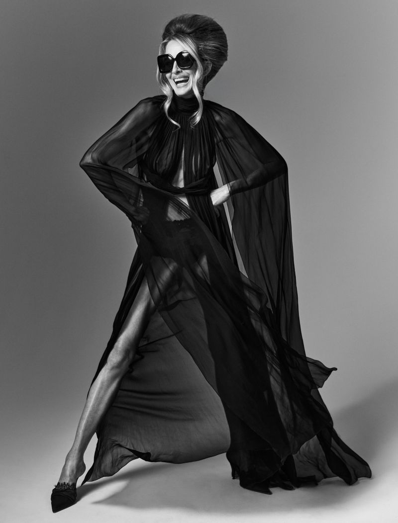 Elegant woman in a black sheer gown and sunglasses poses in a studio. She is smiling with her hand on her hip and leg extended.