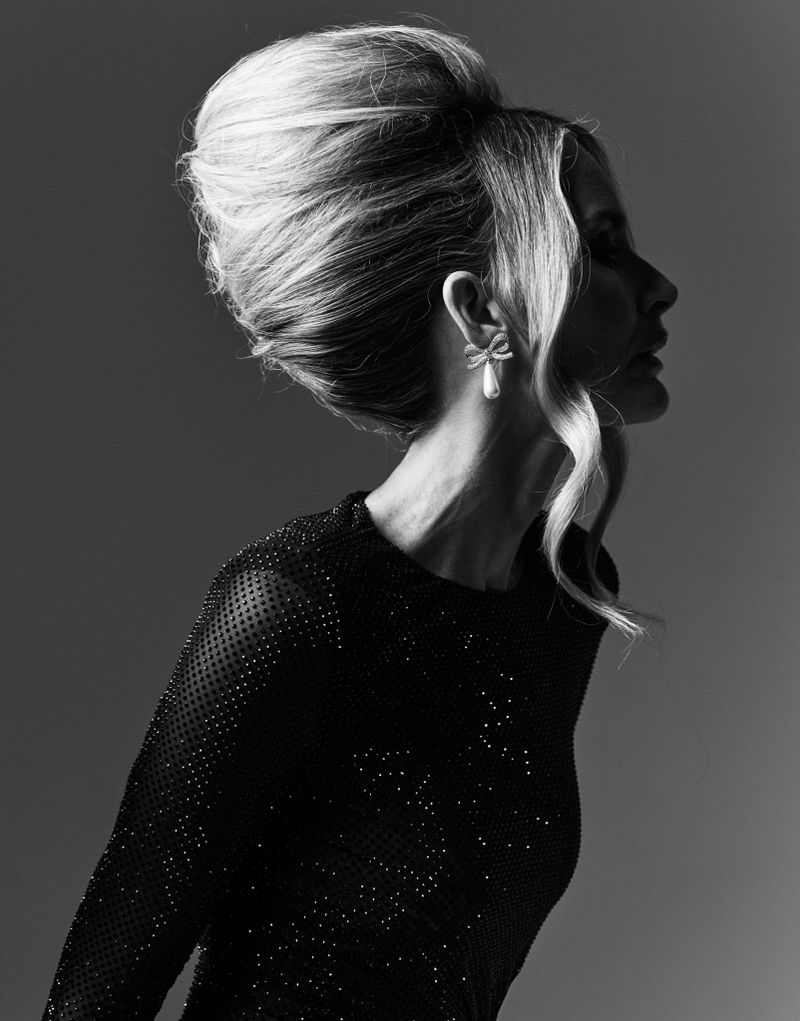 Profile portrait of a woman with blonde hair in a high bouffant updo, wearing a dark beaded top against a graduated gray background.