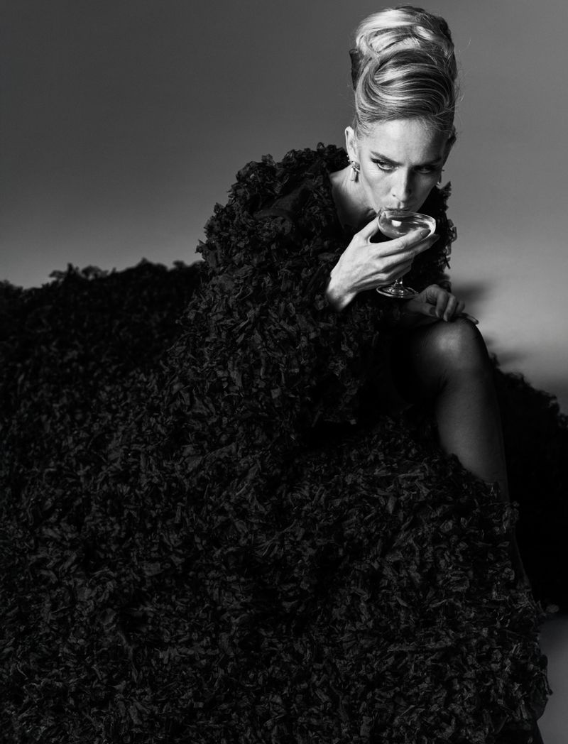 Elegant woman in textured black coat sits and drinks from a stemmed glass in a photo studio against a grey backdrop, black and white.