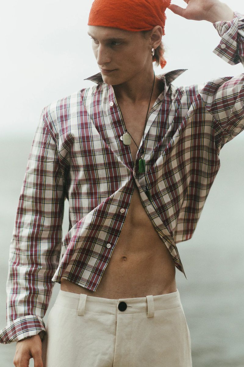 Model Noah Loof wearing an open plaid shirt, cream trousers, and an orange headscarf outdoors against an overcast backdrop.
