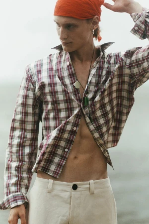 Model Noah Loof wearing an open plaid shirt, cream trousers, and an orange headscarf outdoors against an overcast backdrop.