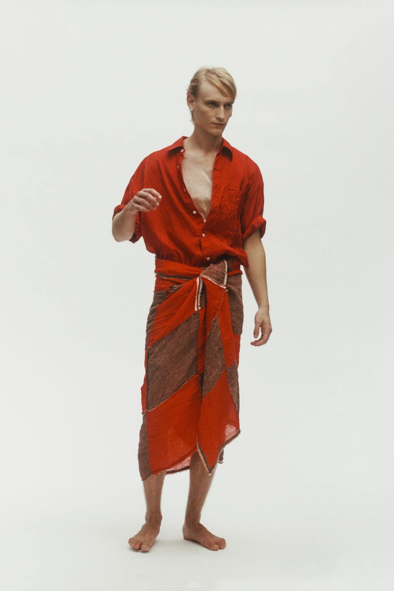 Model Noah Loof wearing a red collared shirt and patterned wrap skirt barefoot against a white backdrop.