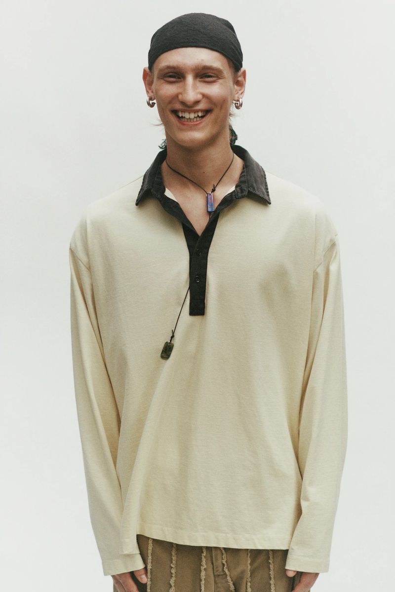 Model Noah Loof smiling in a cream long-sleeve collared polo shirt and black skull cap against a white seamless backdrop.