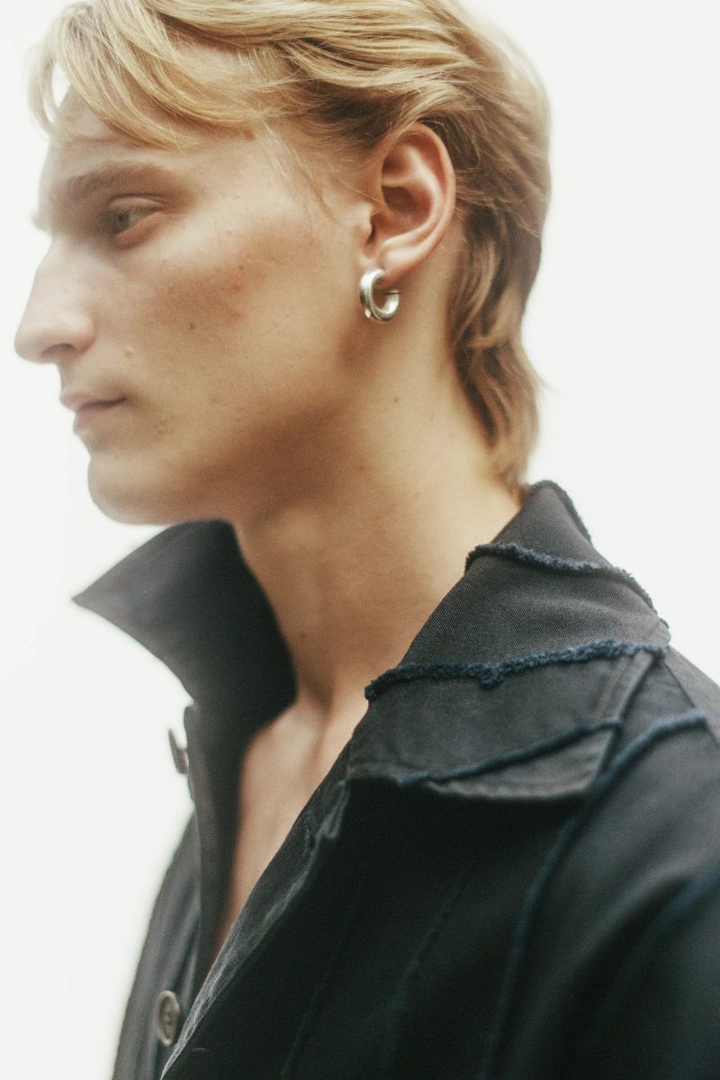 Model Noah Loof in profile wearing a silver hoop earring and a black jacket with raw frayed seams against a white backdrop.