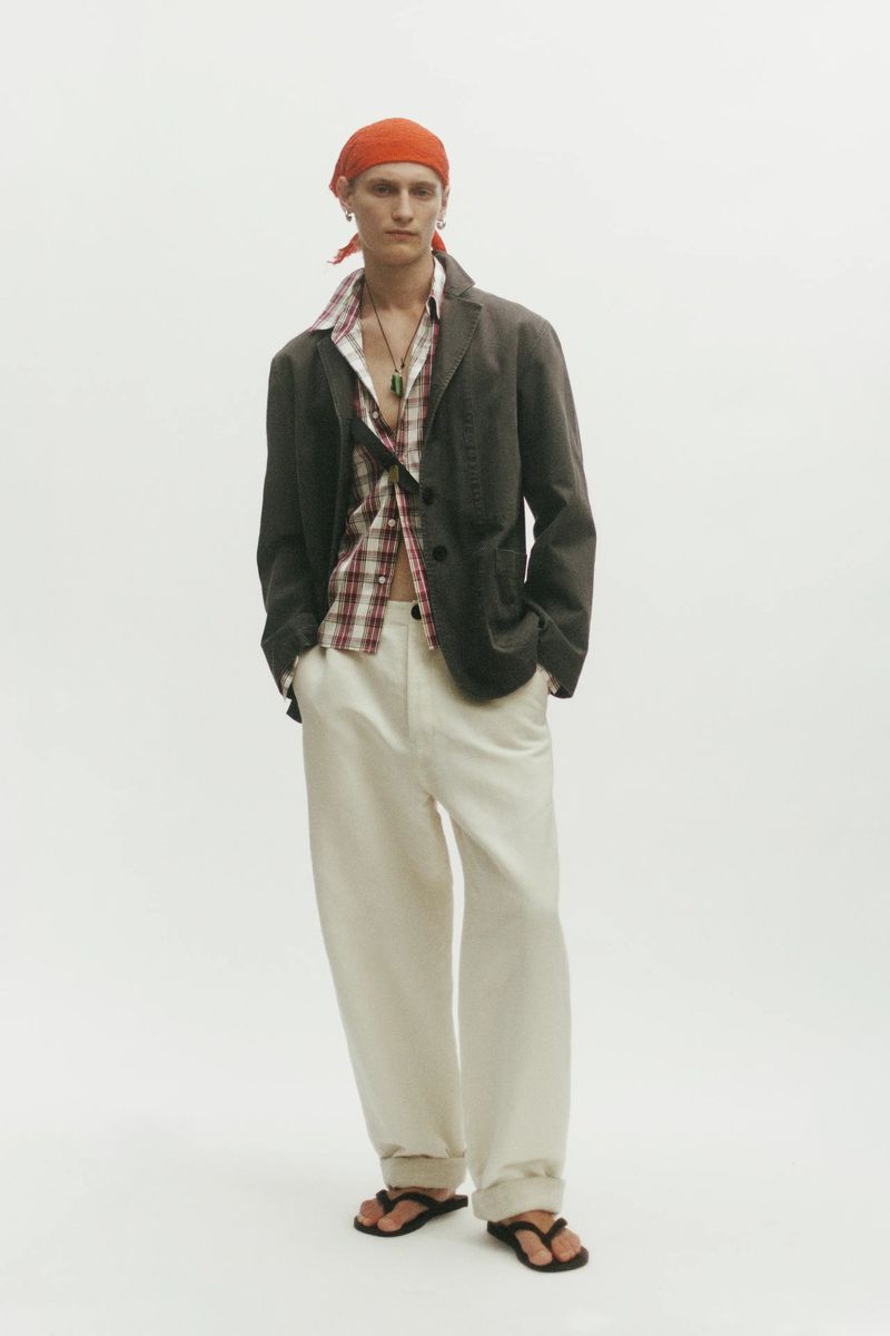 Model Noah Loof wearing a dark blazer, unbuttoned plaid shirt, cream wide-leg trousers, and an orange headscarf on a white backdrop.