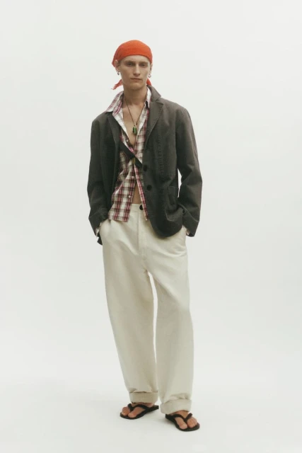 Model Noah Loof wearing a dark blazer, unbuttoned plaid shirt, cream wide-leg trousers, and an orange headscarf on a white backdrop.