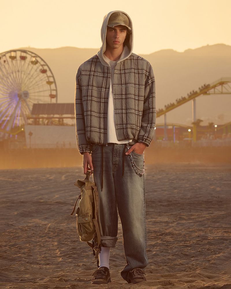 Model wearing a plaid zip jacket, hooded sweatshirt, baggy jeans, and cap on a beach with a Ferris wheel in the background.
