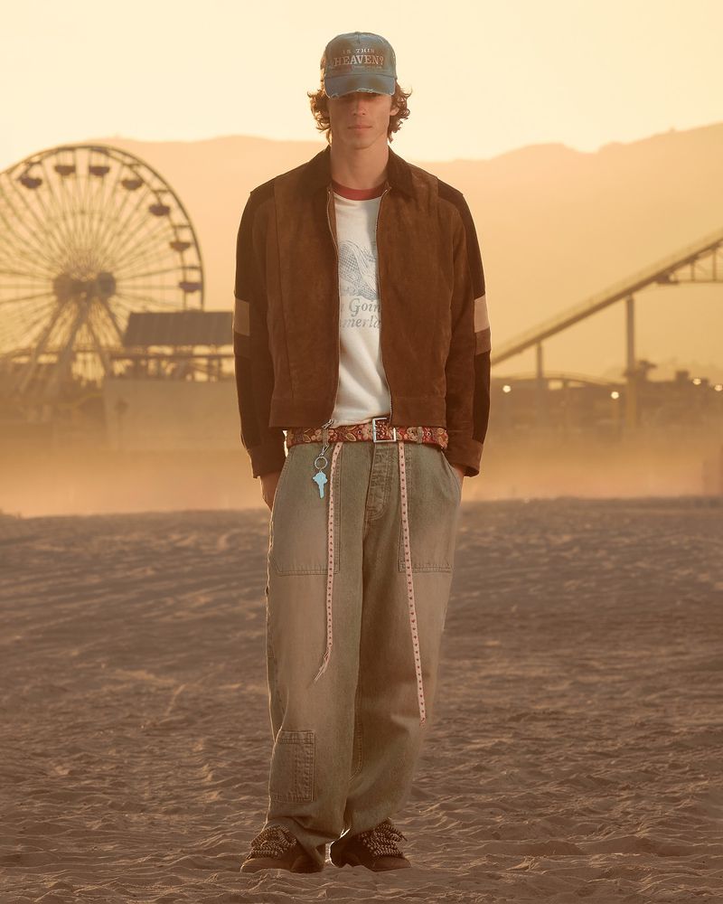 Male model in a brown suede jacket, graphic tee, and baggy pants standing on a hazy beach at sunset with a Ferris wheel behind him.