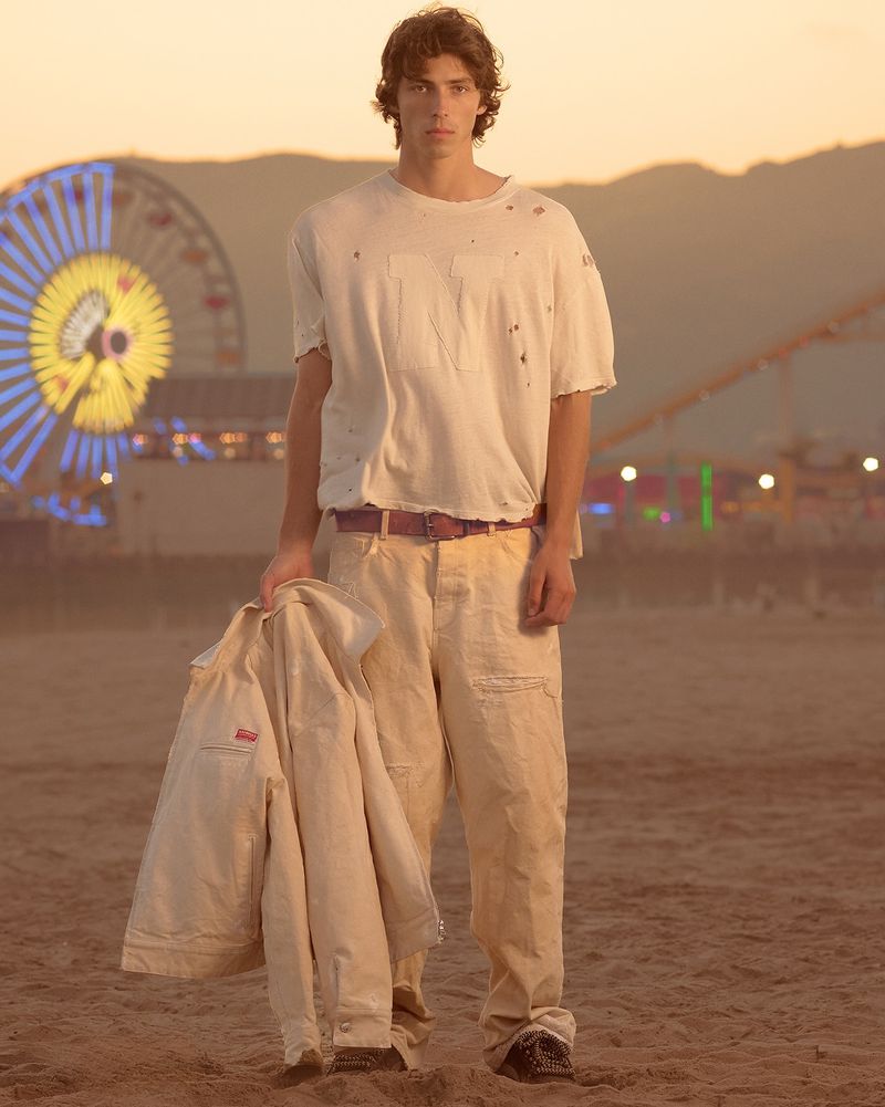 Model standing on a sandy beach at sunset wearing a cream t-shirt and trousers, holding a jacket, with a Ferris wheel in the background