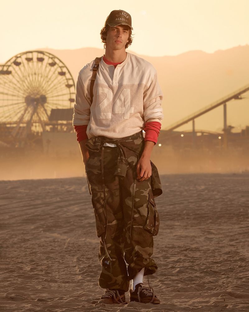 Model wearing camouflage cargo pants and a cream long-sleeve shirt standing on a sandy beach at sunset with a Ferris wheel in the background