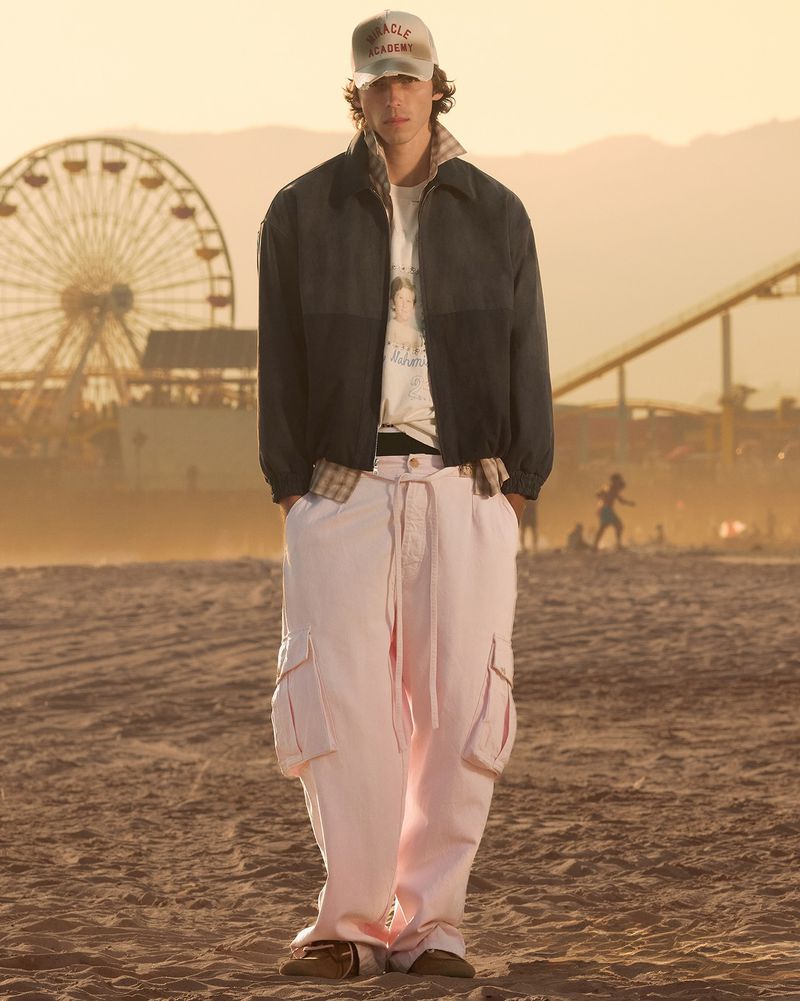 Model wearing a dark jacket and wide-leg pink cargo pants standing on a sandy beach with a distant ferris wheel behind him.
