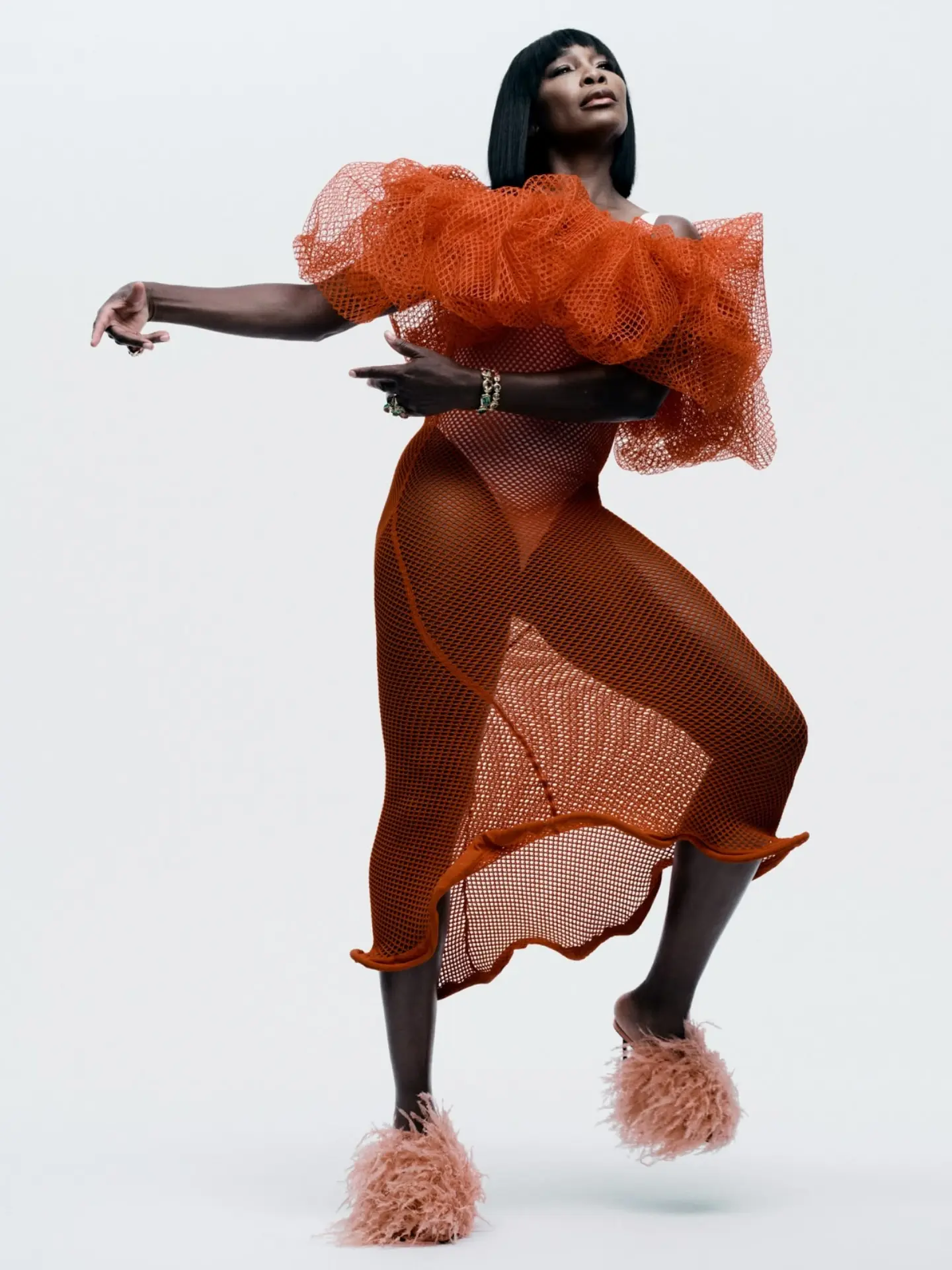 Venus Williams posing in an orange ruffled mesh dress and pink feathered mules against a white seamless studio background.