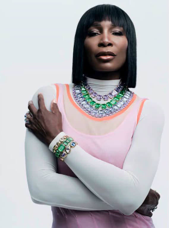 Venus Williams wearing a layered pink dress, statement gemstone necklace, and blunt black bob against a white studio background.