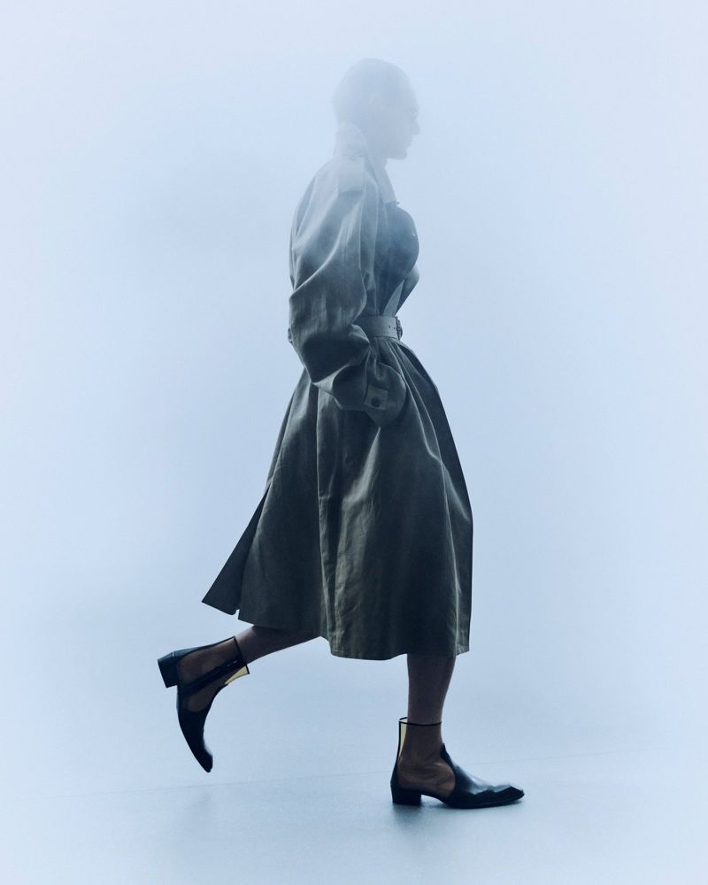 Model walking in profile wearing an oversized belted trench coat and black mesh cutout boots through a misty blue atmospheric haze.