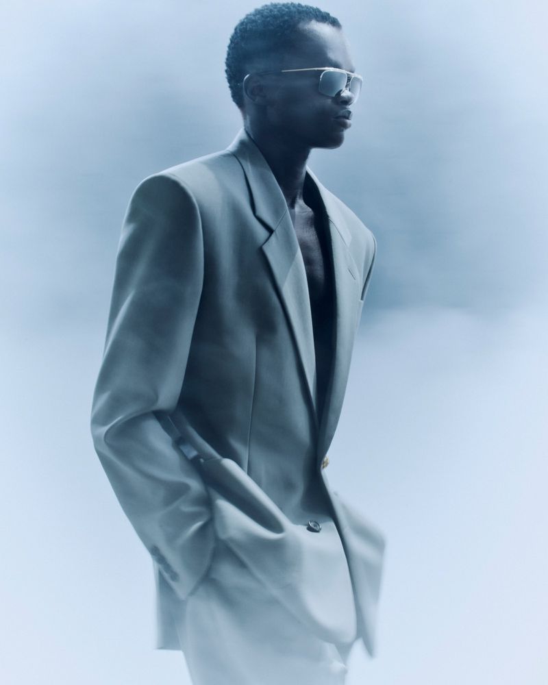 A Black male model wearing an oversized light grey blazer and tinted sunglasses in profile against a hazy blue background.