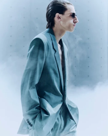 Side profile of a male model wearing sunglasses and an oversized tailored light blue suit surrounded by atmospheric haze.