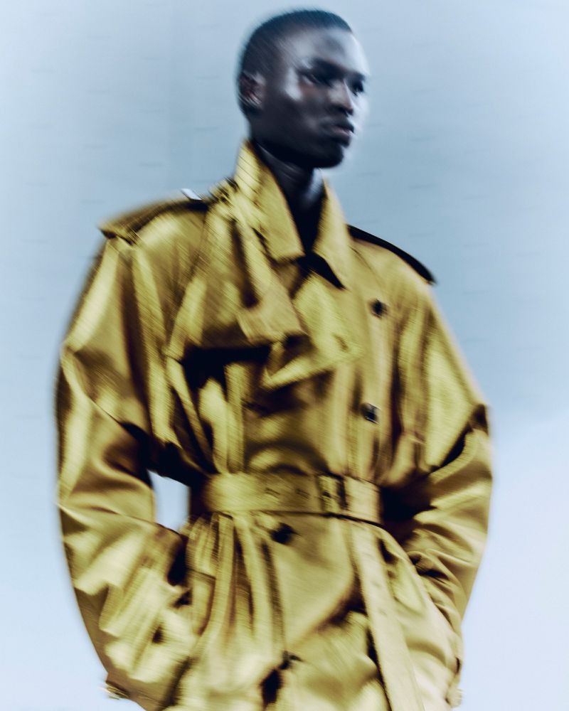 Model wearing a mustard yellow trench coat against a plain background in a motion-blurred fashion photograph.