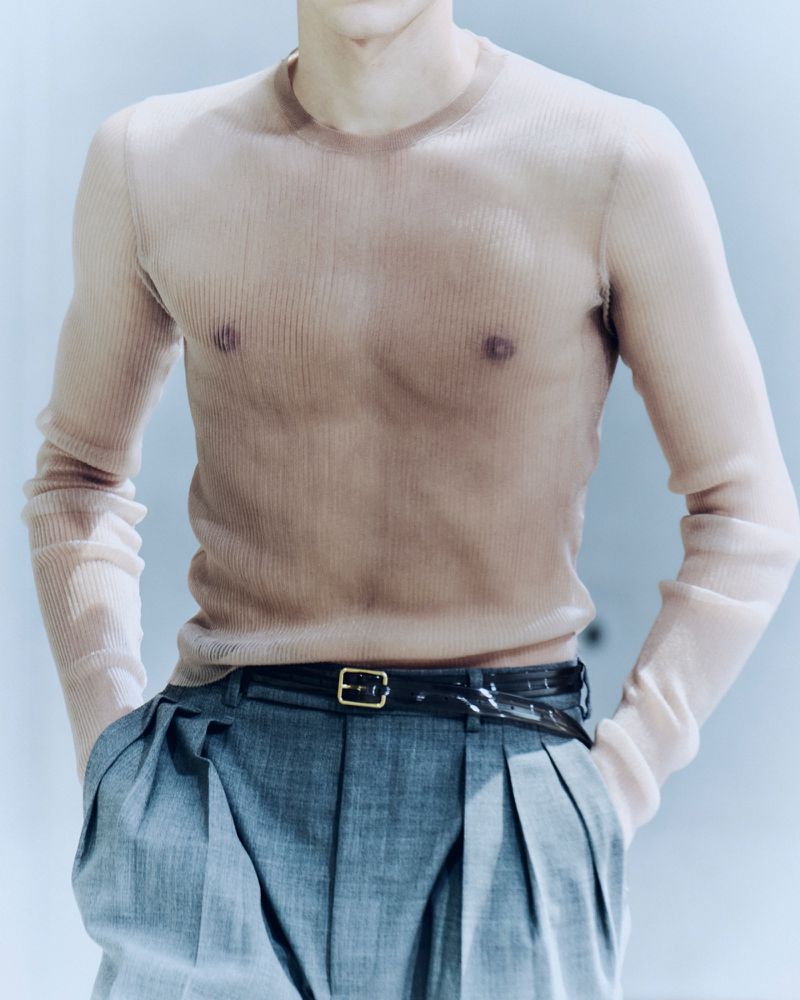 Close-up of a model wearing a sheer beige ribbed long-sleeve top and pleated grey trousers with a black belt against a pale backdrop.