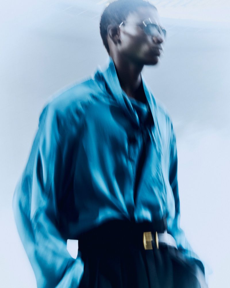 Motion-blurred portrait of a Black male model wearing a bright blue silk shirt and black trousers with a gold belt buckle.