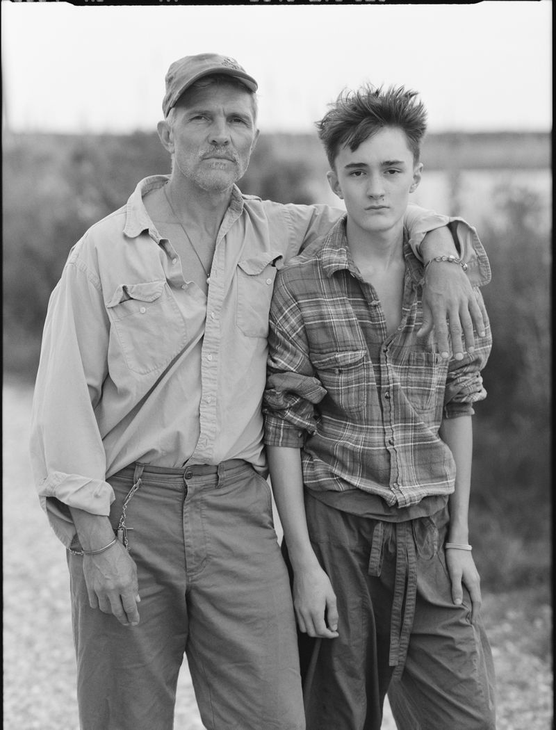 Black and white portrait of an older man with a cap and younger person in a plaid shirt standing outdoors together.