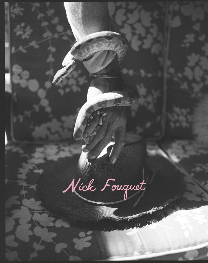 Black and white photo of a snake wrapped around a hand resting on a wide-brimmed hat on a floral patterned chair.