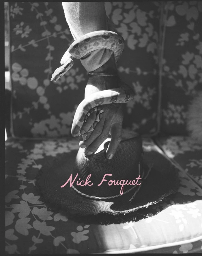Black and white photo of a snake wrapped around a hand resting on a wide-brimmed hat on a floral patterned chair.
