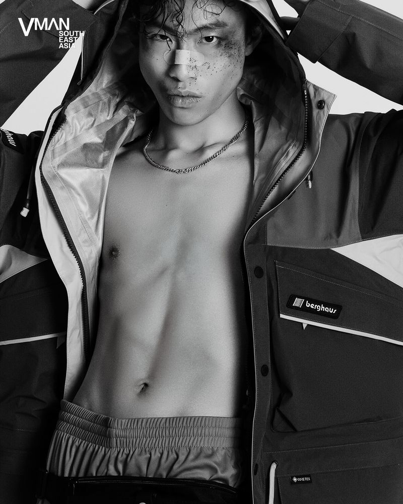East Asian male model posing shirtless in an open hooded technical jacket with face makeup and nose tape against a plain backdrop.