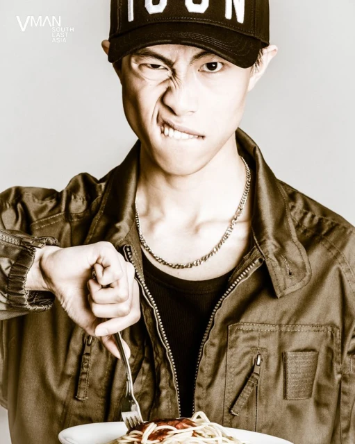 Sepia portrait of a young adult man eating pasta with a fork, wearing a baseball cap and jacket, in an indoor setting.