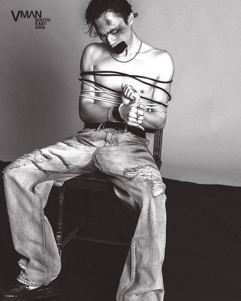 Monochrome studio shot of a topless man tied to a wooden chair with cables and tape over his mouth, wearing ripped jeans and jewelry.