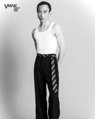 Black and white portrait of a lean adult male in a white tank top and black pants, with a striped tie as a belt, posing in a studio.