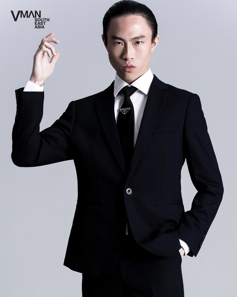Portrait of an East Asian man in a black suit and tie, hand raised slightly, against a neutral background in a studio setting.