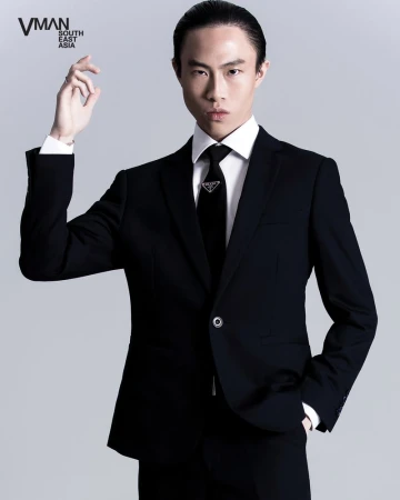 Portrait of an East Asian man in a black suit and tie, hand raised slightly, against a neutral background in a studio setting.