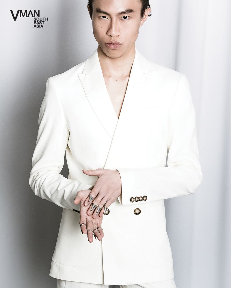 Fashion portrait of an Asian male model in a white double-breasted suit, striking a pose against a soft grey studio background with a clean aesthetic.
